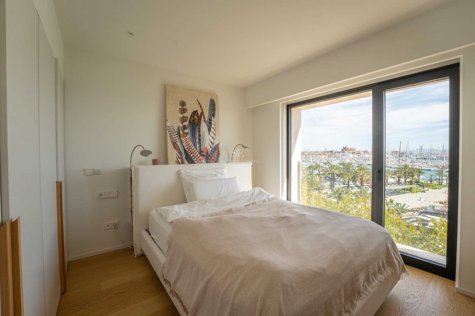 Chambre moderne avec lit blanc et grande fenêtre offrant une vue sur une marina ensoleillée avec des palmiers.