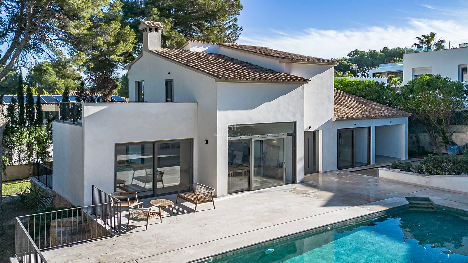 Villa blanca moderna con techo de tejas, puertas de cristal y terraza de piedra junto a una piscina turquesa.