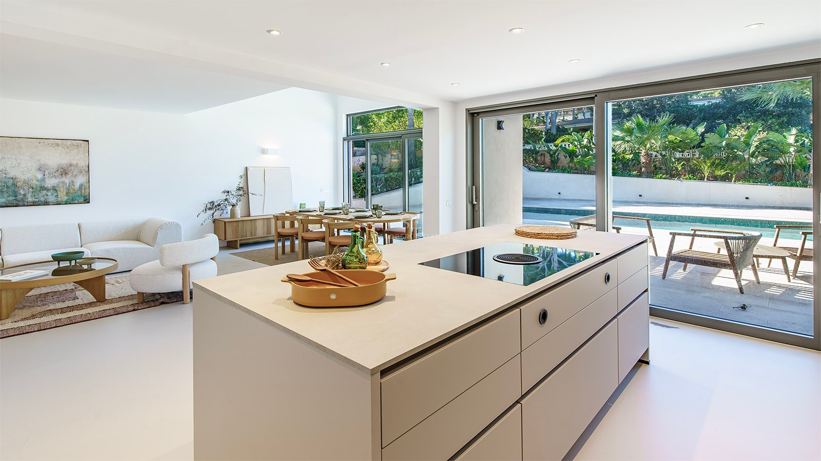 Interior luminoso y moderno con isla de cocina, comedor, sofás y grandes ventanales que dan a una piscina.