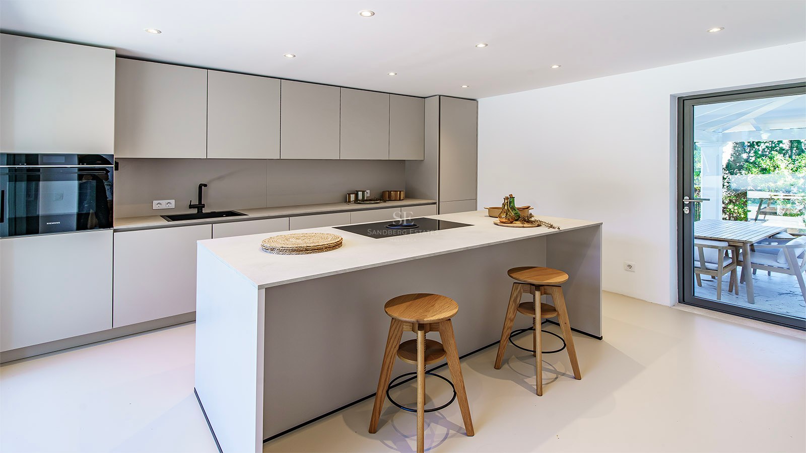 Elegante cocina gris claro con una gran isla, taburetes de madera y puerta de cristal hacia la terraza.