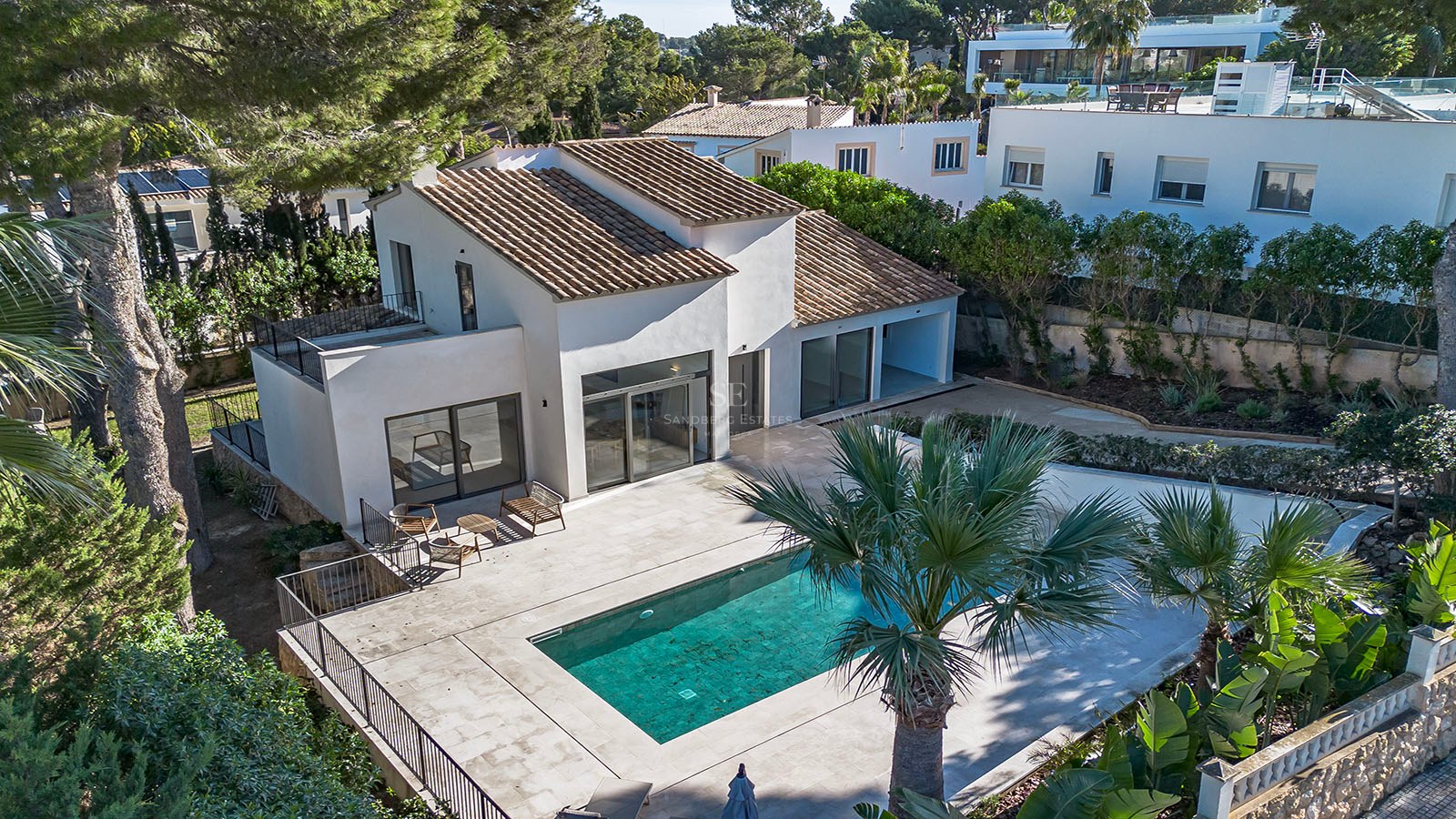 Vista aérea de una villa blanca con tejado de teja, piscina turquesa y amplia terraza rodeada de pinos y palmeras.