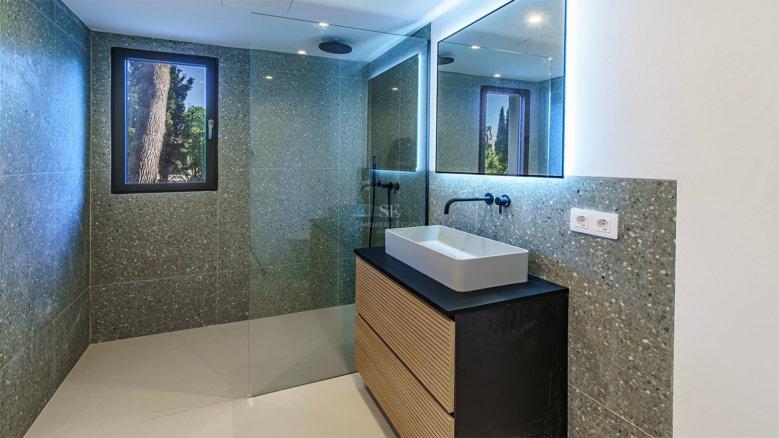 Baño moderno con paredes de terrazo gris, ducha de cristal, grifería negra y mueble de madera con lavabo blanco.