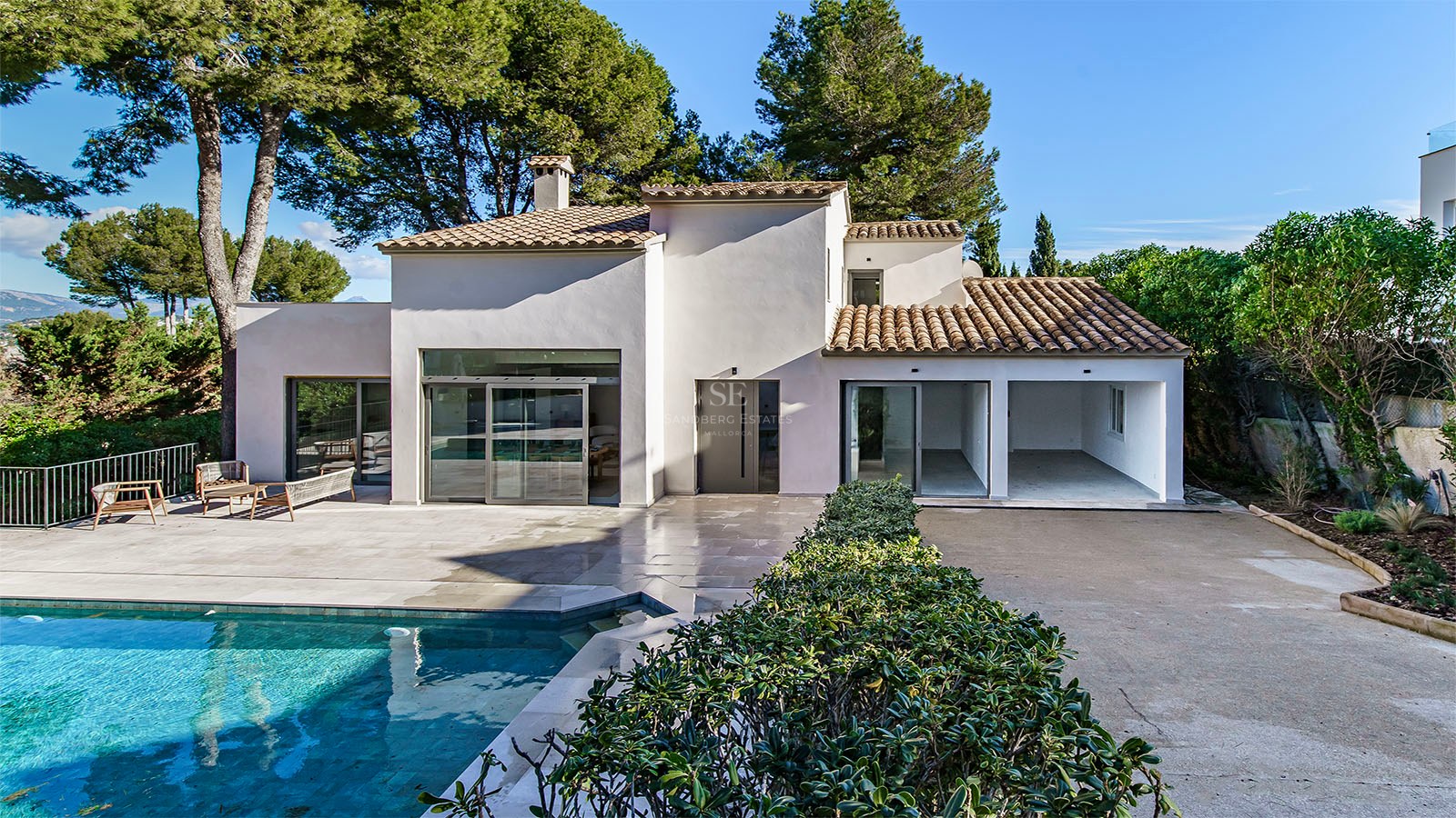 Villa blanca moderna con piscina turquesa, terraza de piedra y pinos bajo un cielo azul despejado.