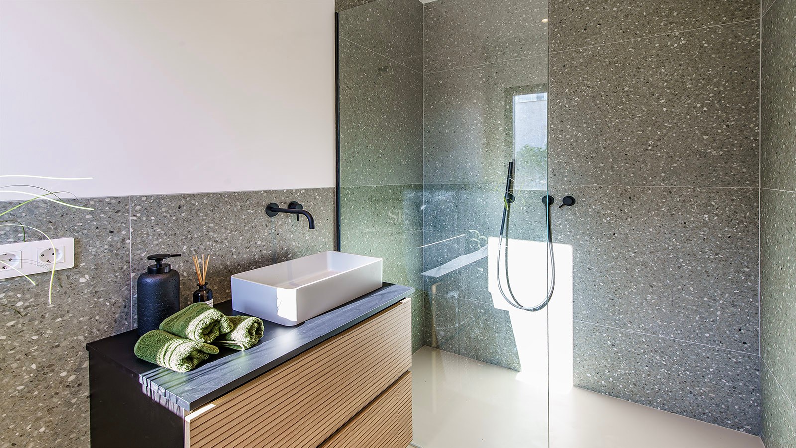 Baño moderno con azulejos grises moteados, lavabo blanco sobre mueble de madera y ducha con mampara de cristal.