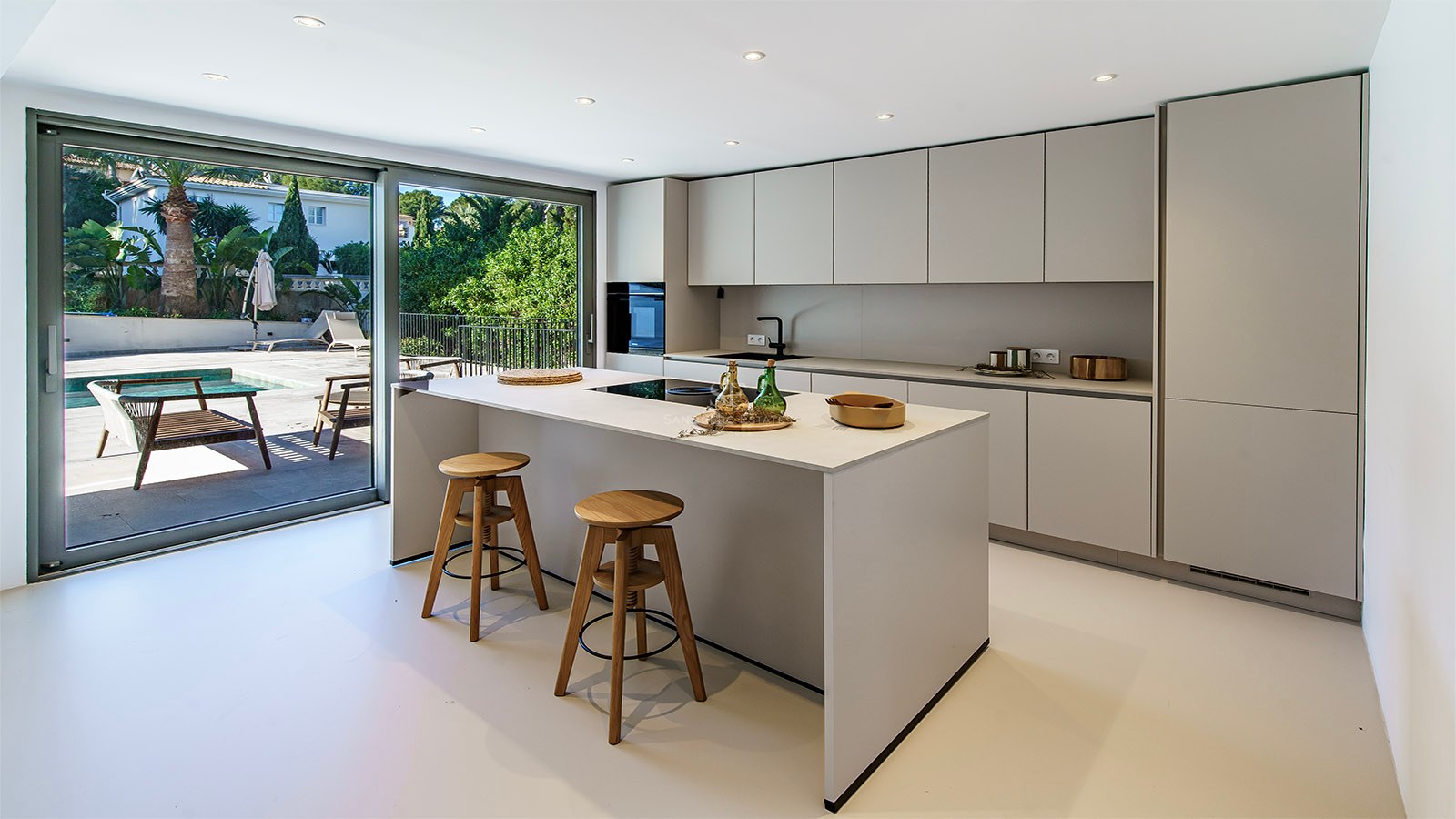 Cocina moderna blanca con isla central, dos taburetes de madera y grandes puertas correderas de cristal hacia la piscina.