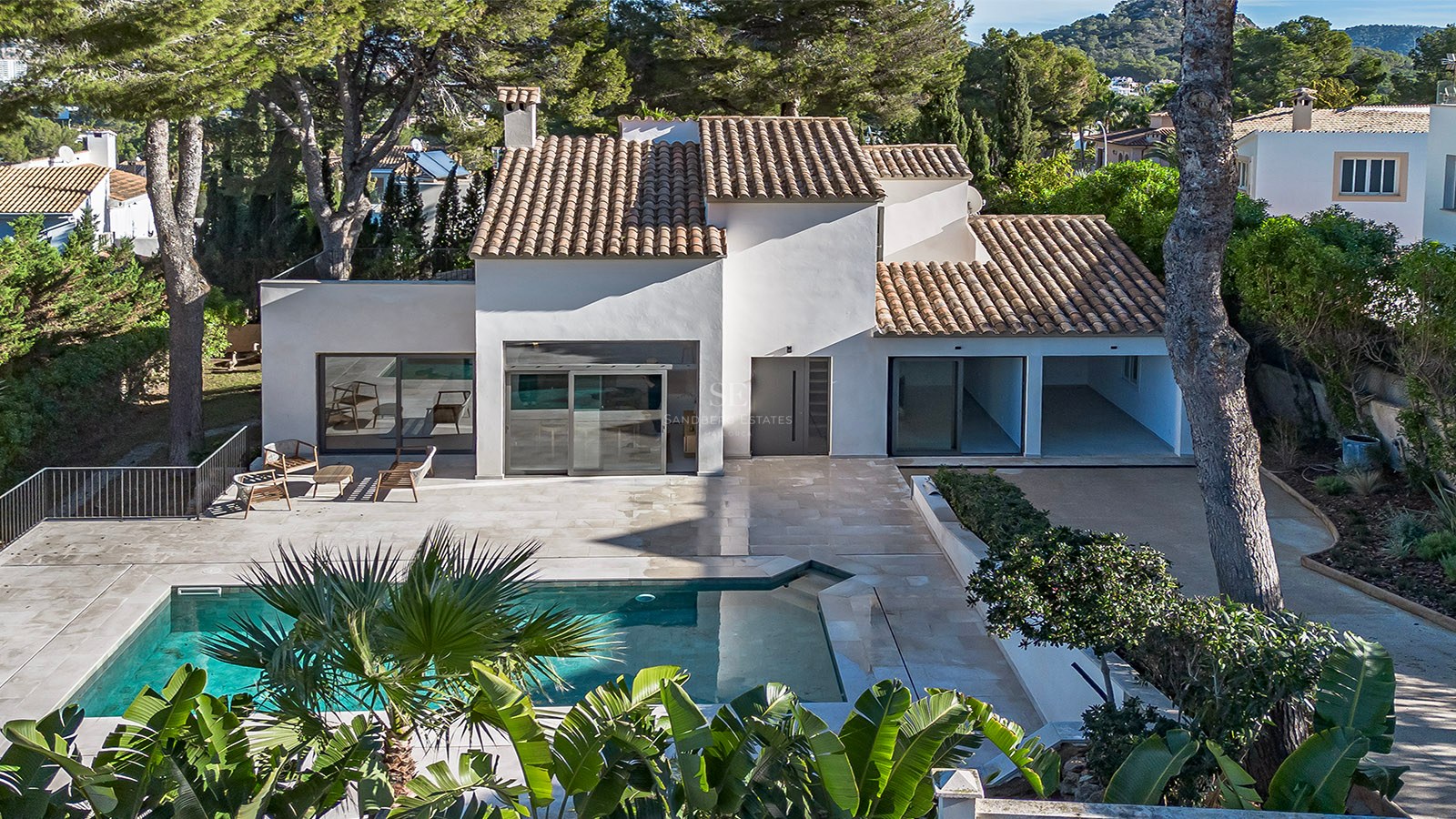 Vista aérea de una villa blanca con techo de teja, piscina turquesa y terraza de piedra rodeada de pinos.