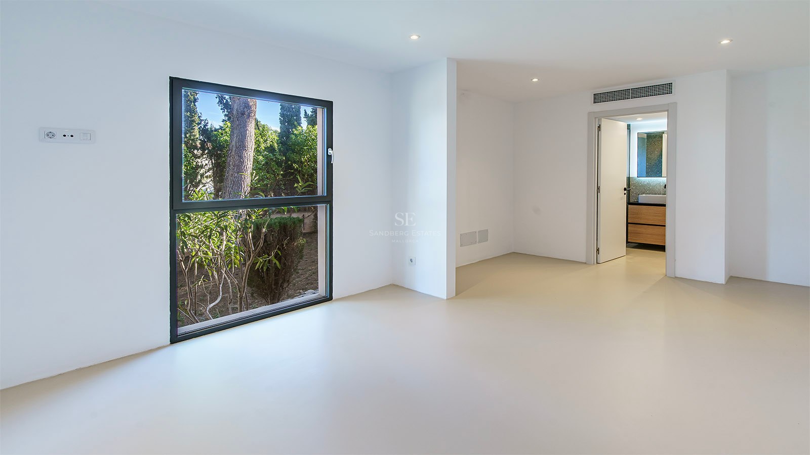 Dormitorio minimalista vacío con paredes blancas, suelo beige, ventana al jardín y puerta a un baño en suite.