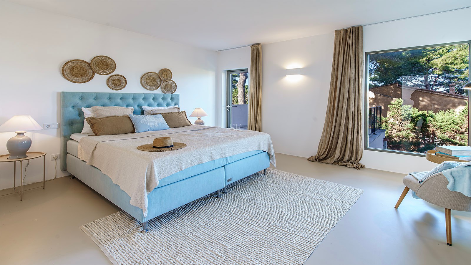 Habitación luminosa con cama azul claro, alfombra de lana y gran ventanal con vistas a árboles verdes.