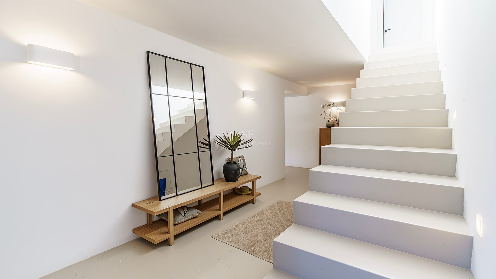 Pasillo minimalista luminoso con escaleras blancas, un gran espejo de marco negro, banco de madera e iluminación cálida.