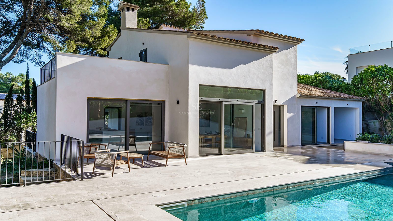 Una gran piscina turquesa frente a una villa blanca moderna con terraza de piedra y muebles de exterior.