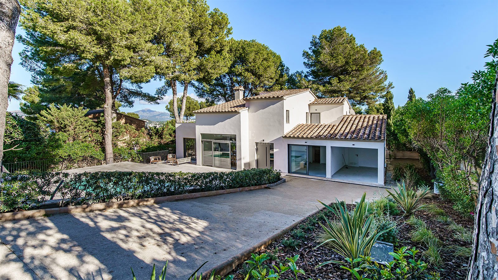 Una villa blanca moderna de dos plantas con tejas de terracota, rodeada de altos pinos y una zona de piscina.