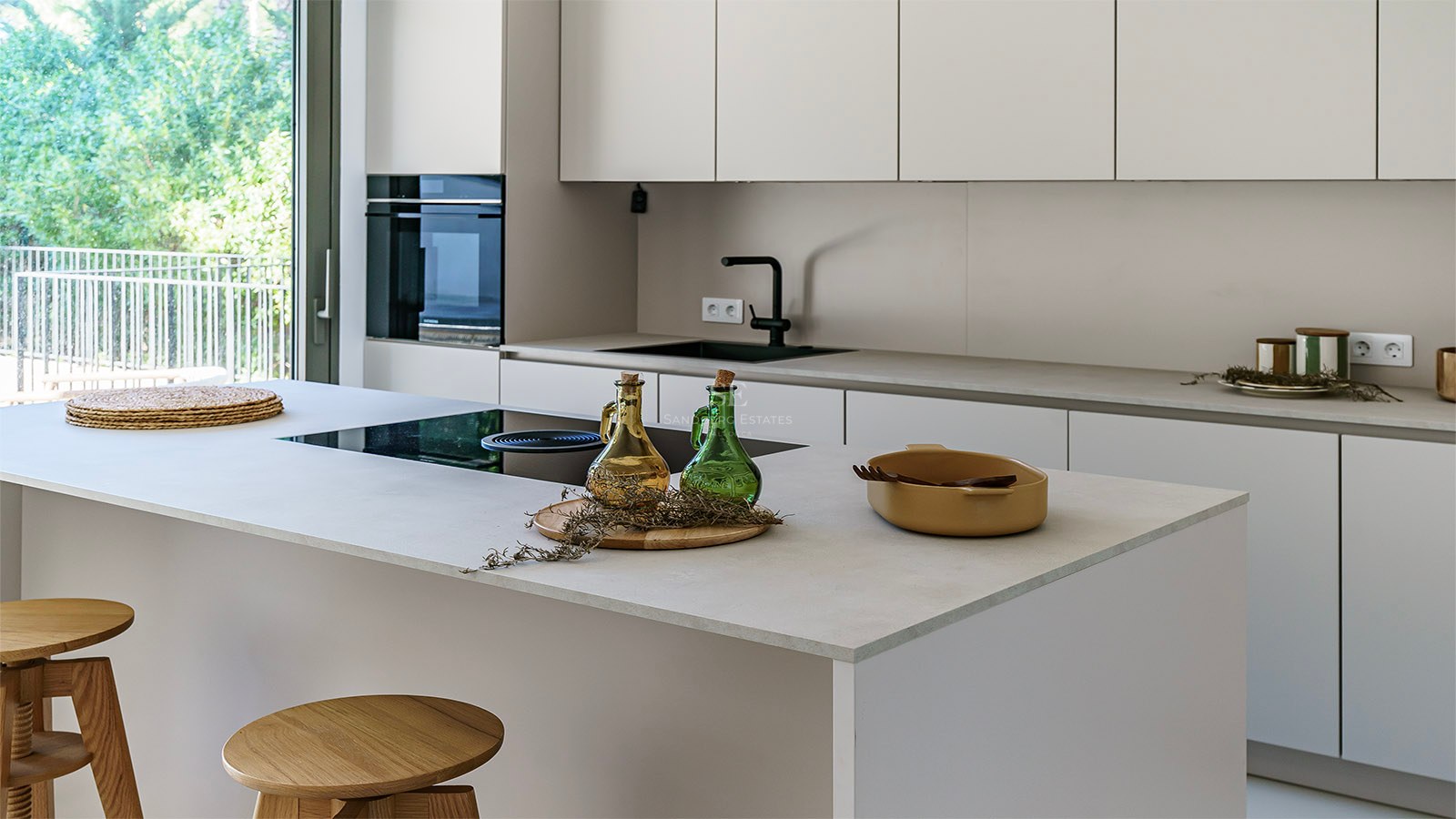 Cocina blanca minimalista con isla central, taburetes de madera y ventanal con vistas a un jardín frondoso.