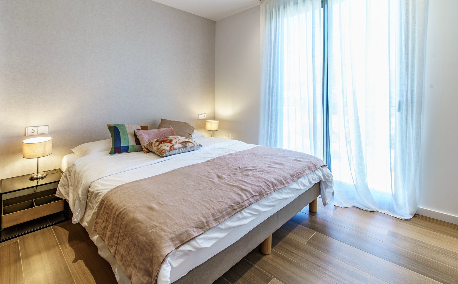 Chambre lumineuse avec un grand lit, des coussins décoratifs, des lampes de chevet et de grandes fenêtres avec voilages.