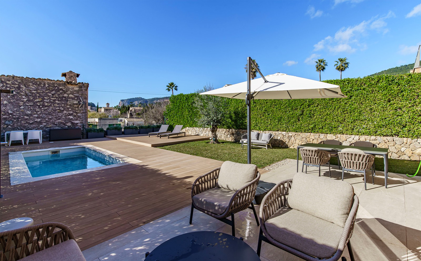 Piscine moderne avec terrasse en bois, mobilier d'extérieur élégant et haies verdoyantes sous un ciel bleu.