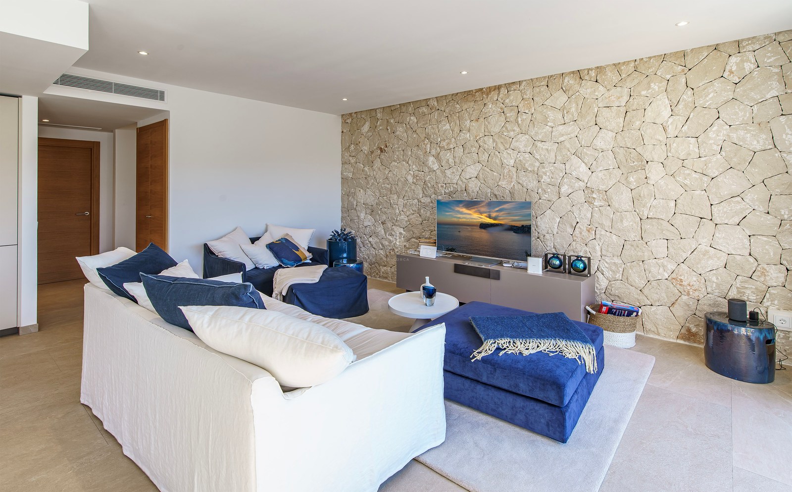 Salon contemporain avec canapé blanc, poufs bleu marine et mur en pierre naturelle texturée avec télévision.
