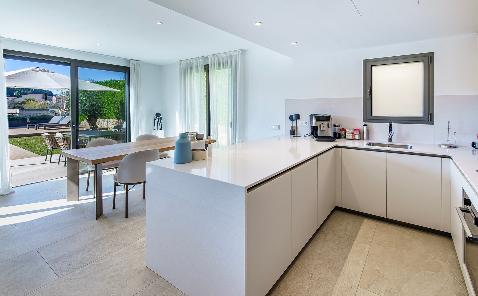 Cuisine minimaliste blanche avec îlot central, table à manger en bois et baies vitrées ouvrant sur un jardin.
