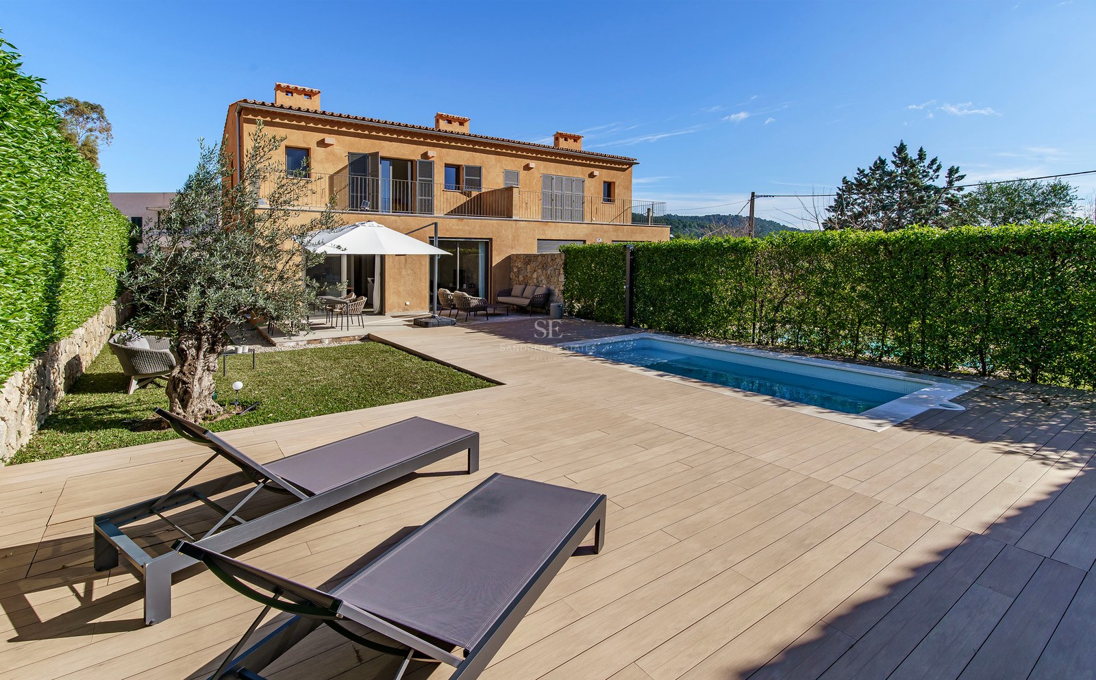 Piscine moderne et terrasse en bois avec transats à côté d'une villa en terre cuite sous un ciel bleu clair.