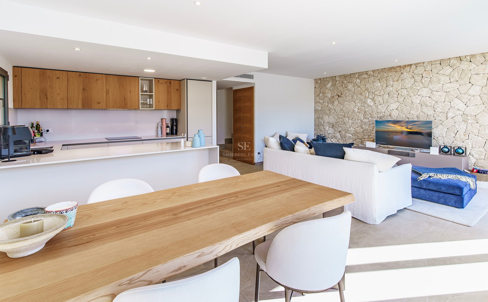 Pièce lumineuse à aire ouverte avec table à manger en bois, cuisine moderne, canapé blanc et mur en pierre naturelle.