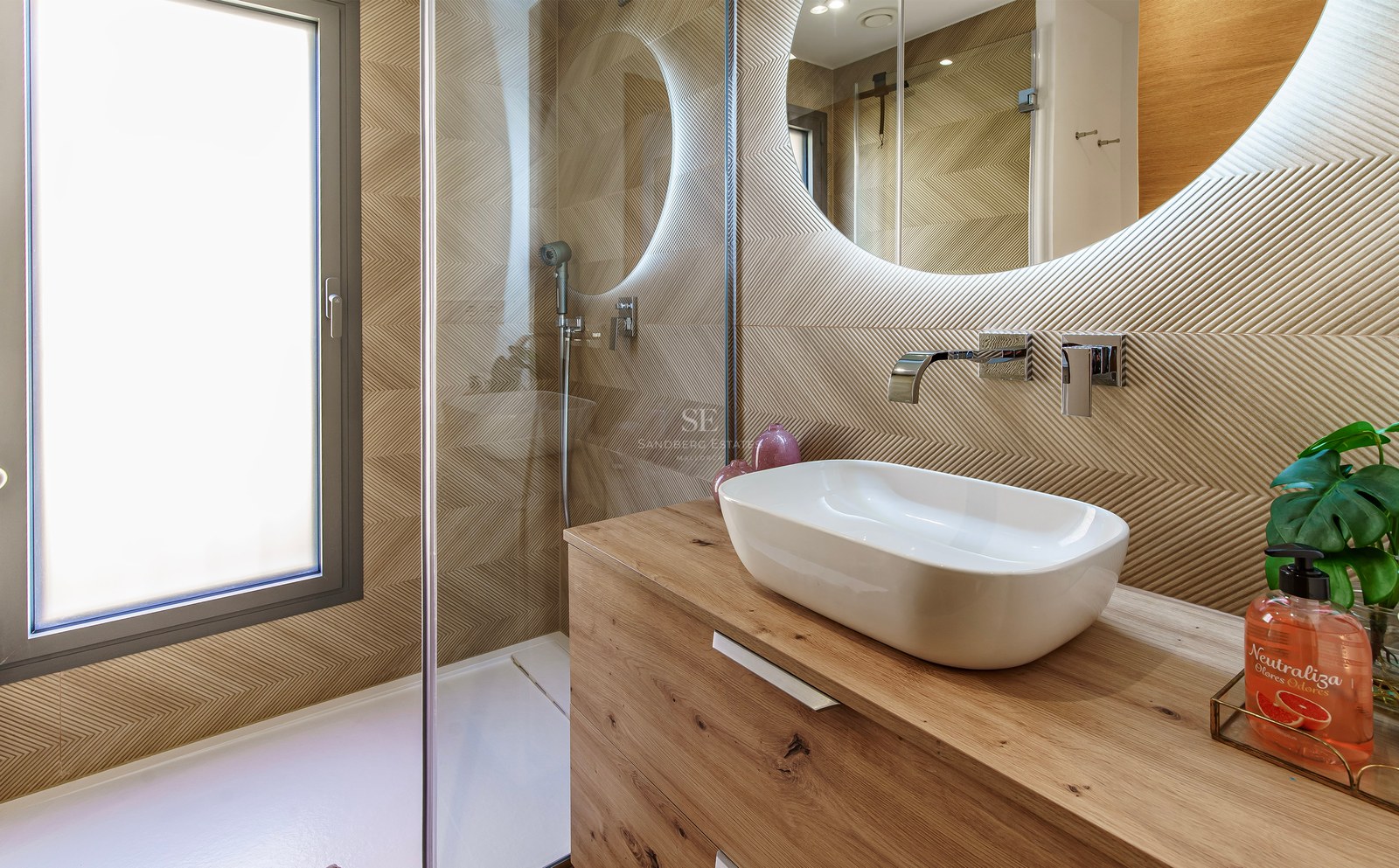 Salle de bain avec vasque à poser sur bois, miroir rond rétroéclairé et paroi de douche en verre.