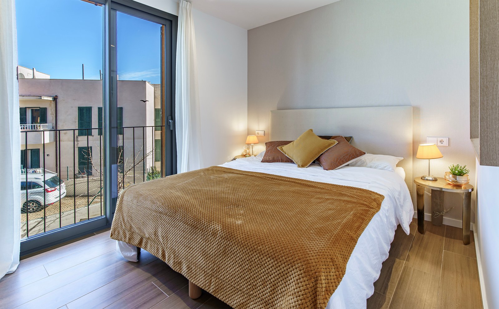Chambre lumineuse avec un lit double, un jeté jaune ocre, une grande fenêtre et un balcon.