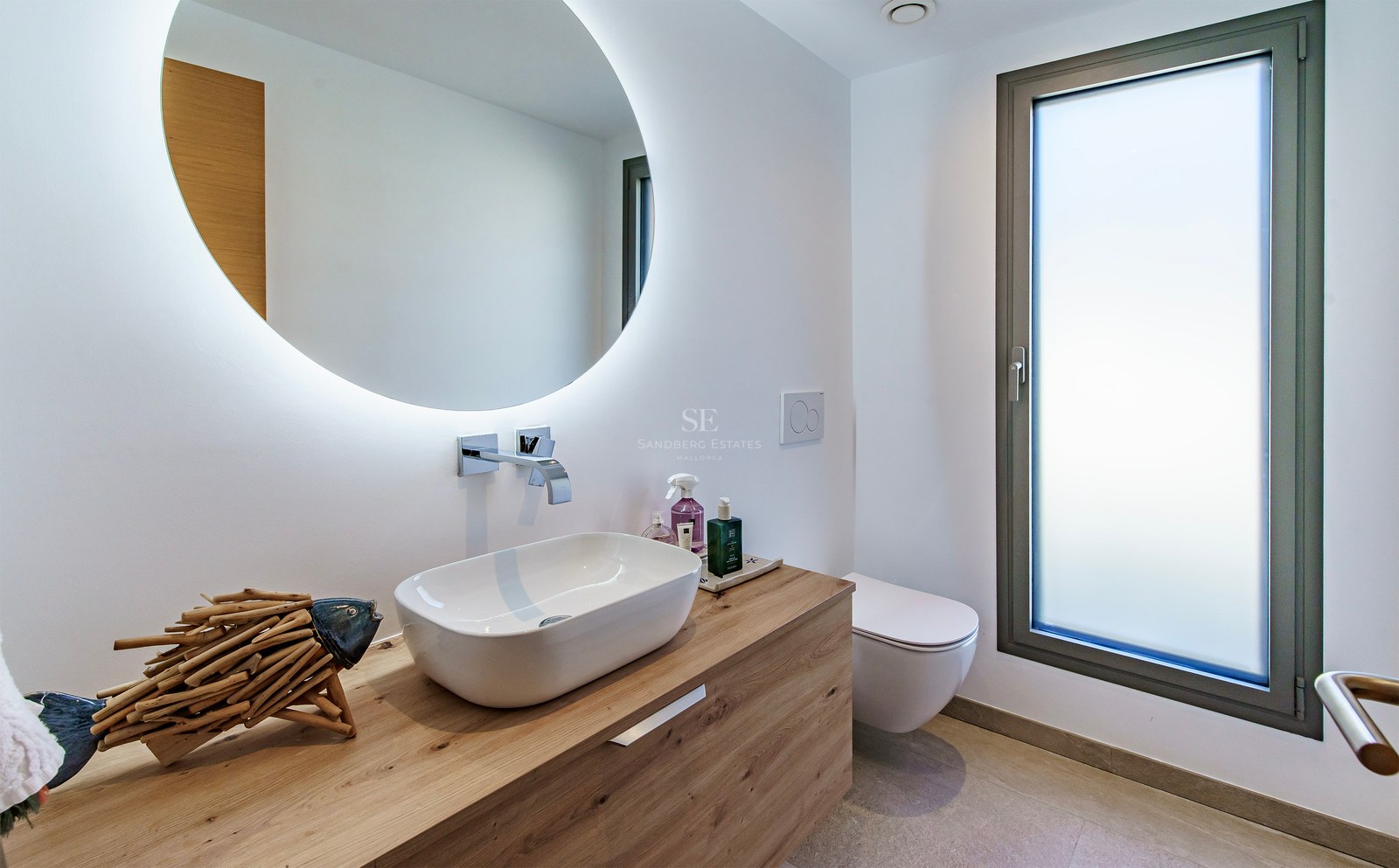 Salle de bain moderne avec vasque sur meuble en bois, miroir rond éclairé et fenêtre en verre dépoli.
