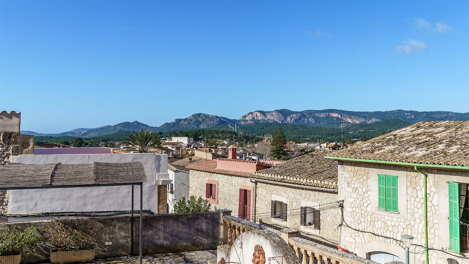 Utsikt från en stenterrass över terrakottatak och gröna berg under en klarblå himmel på Mallorca.