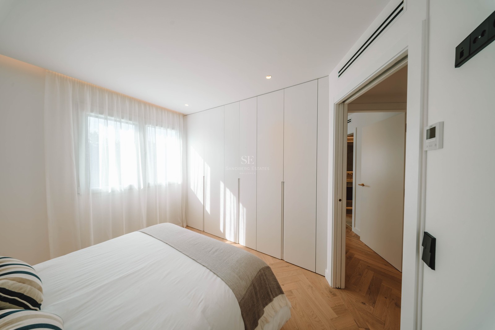 Bright bedroom featuring herringbone wood floors, floor-to-ceiling white wardrobes, a bed with white linens, and sheer curtains.