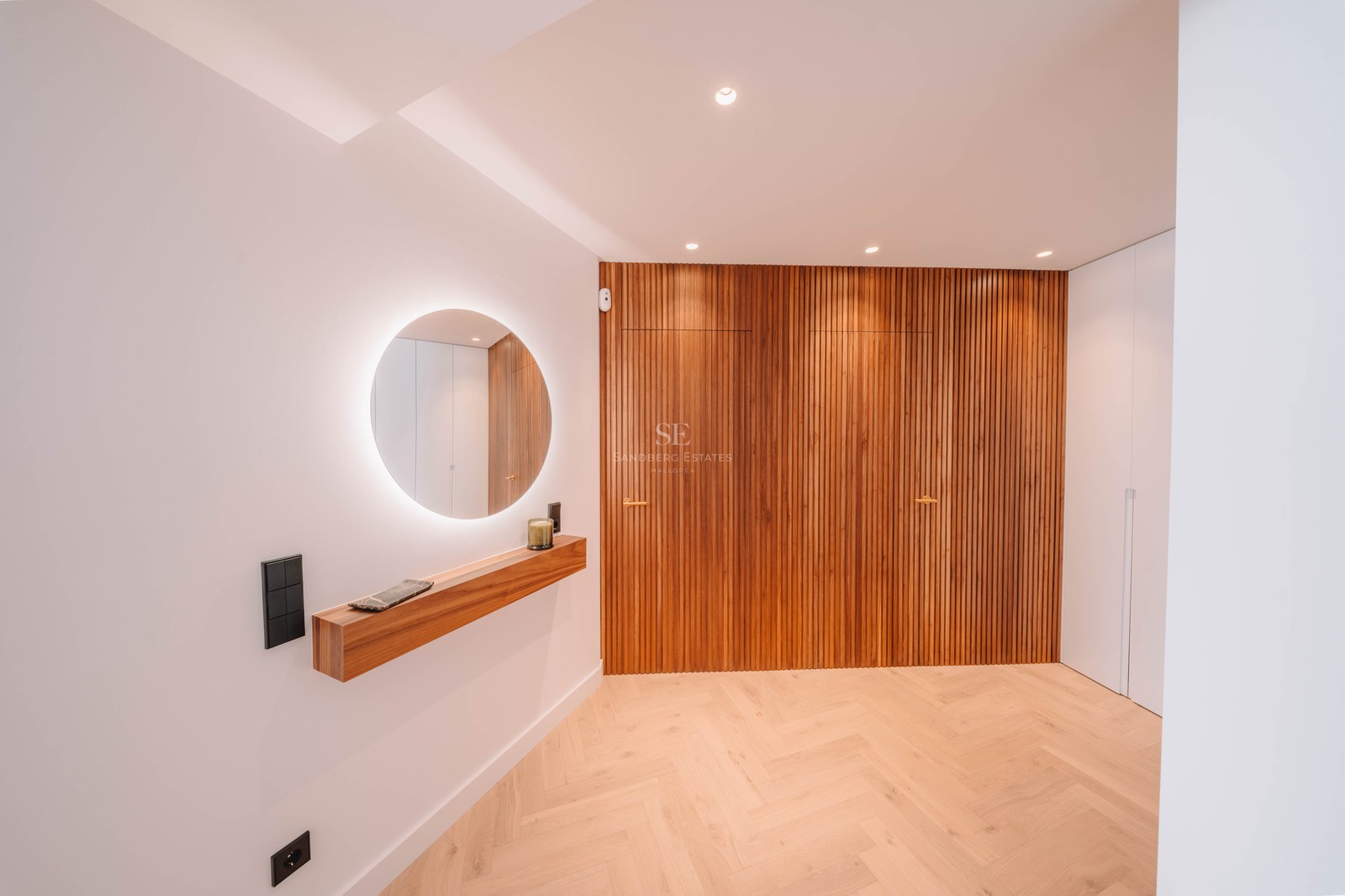 Minimalist hallway featuring a backlit circular mirror, wooden floating shelf, and vertical wood slat wall.