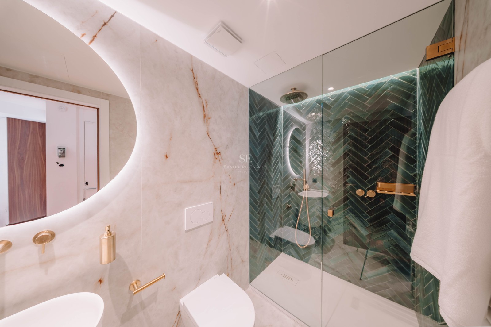Modern bathroom with white marble walls, a round backlit mirror, and a walk-in shower with green herringbone tiles.