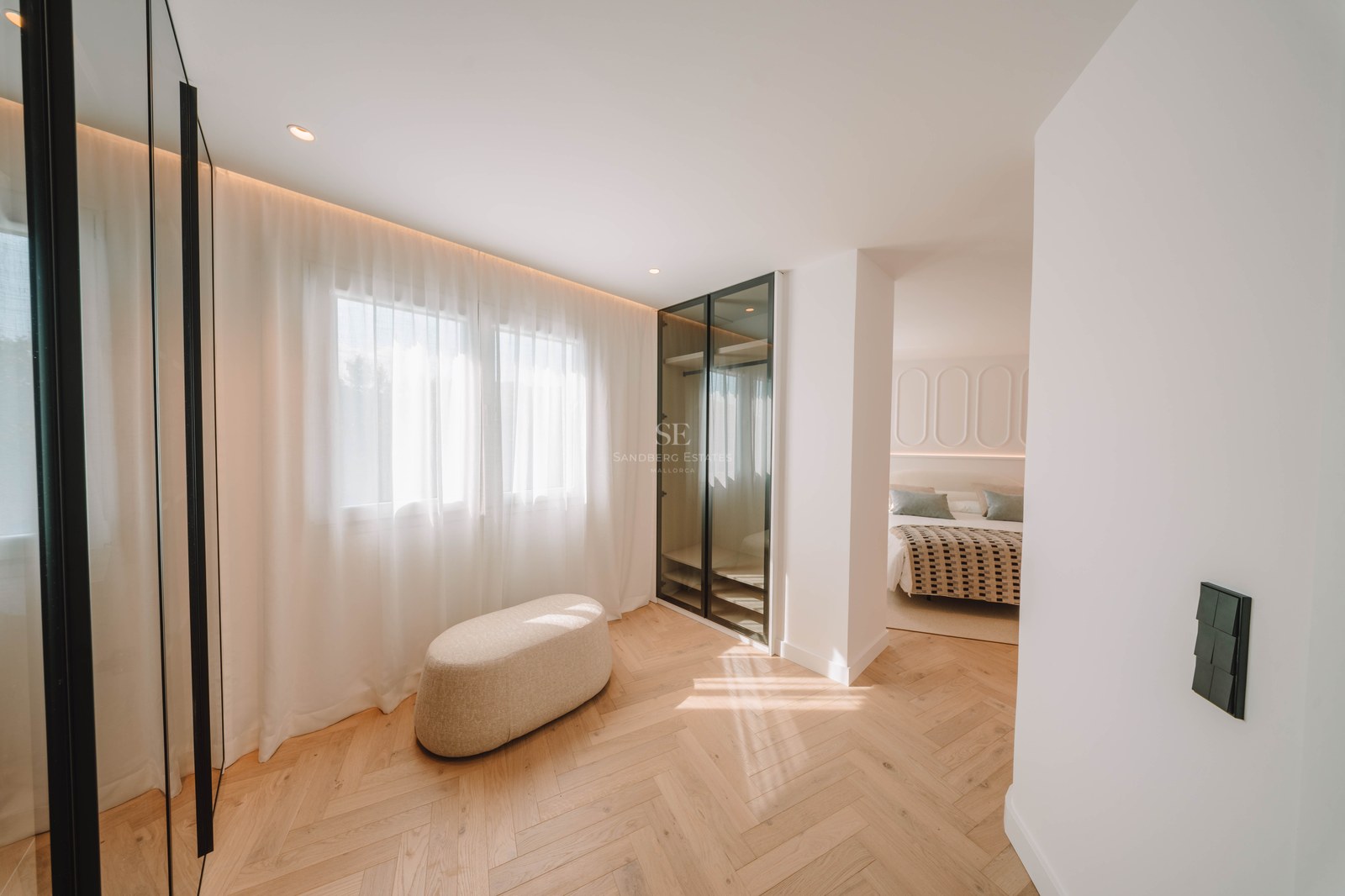 Bright dressing area with herringbone wood flooring, a beige ottoman, and glass wardrobe leading to a bedroom.