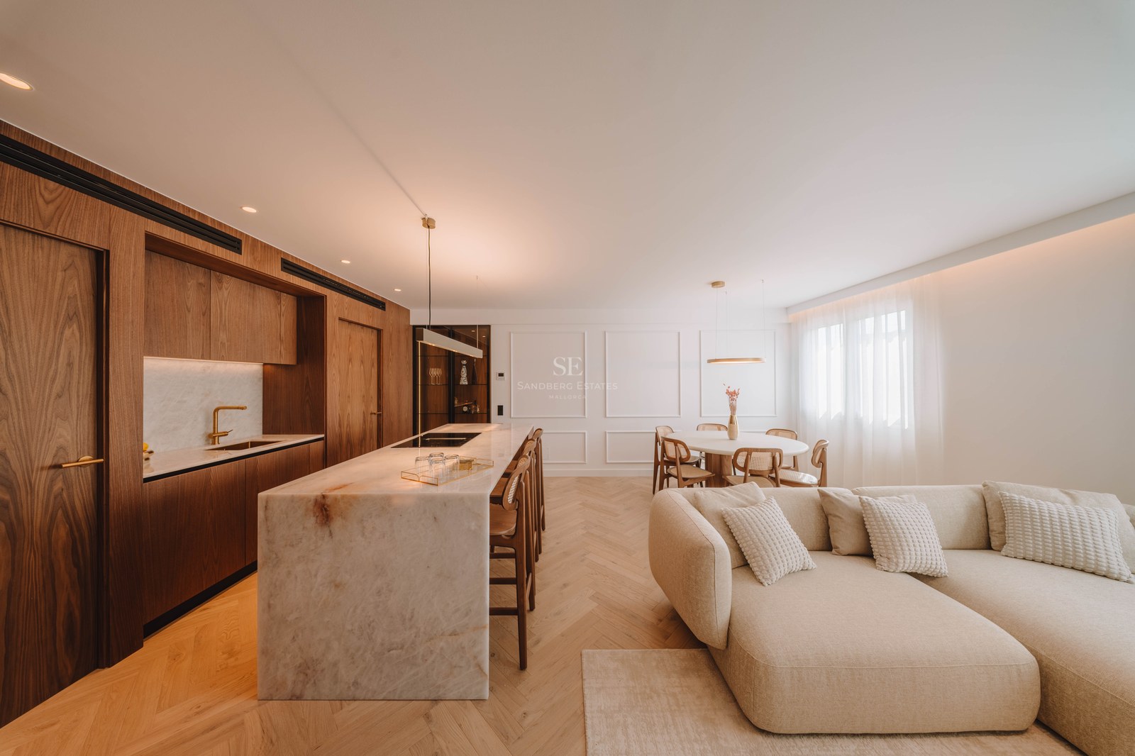 Modern open-plan room with a marble kitchen island, wood cabinets, herringbone floors, and a large cream sectional sofa.