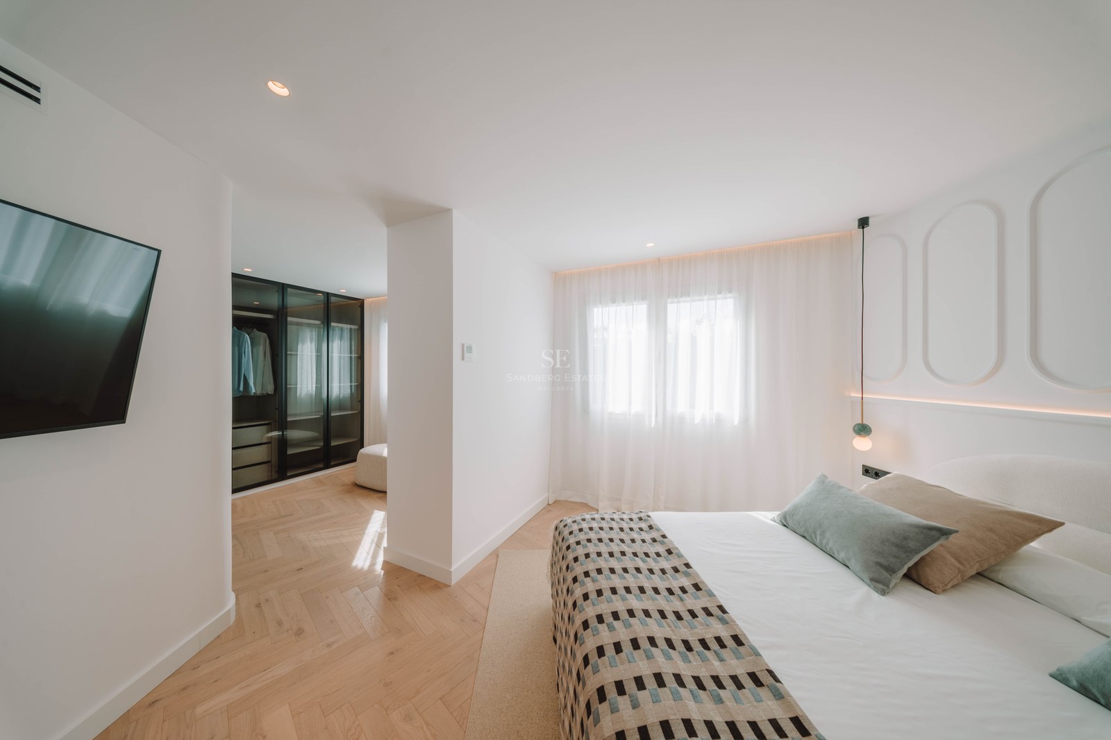 Bright master bedroom featuring herringbone wood floors, a large bed with pillows, and a glass-front walk-in closet.