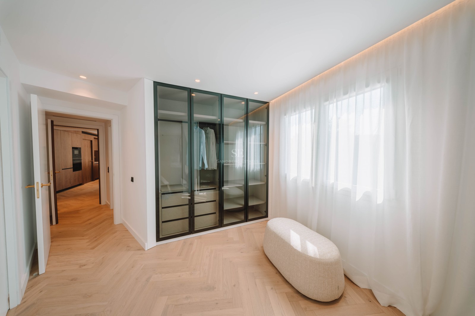 Modern walk-in closet with herringbone wood floors, glass wardrobe, and a central ottoman next to a window.