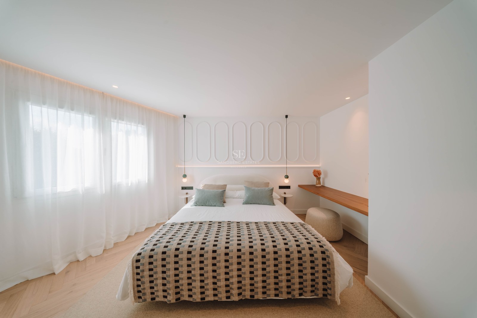 A bright, modern bedroom featuring a patterned bedspread, white paneled headboard wall, and herringbone wood flooring.