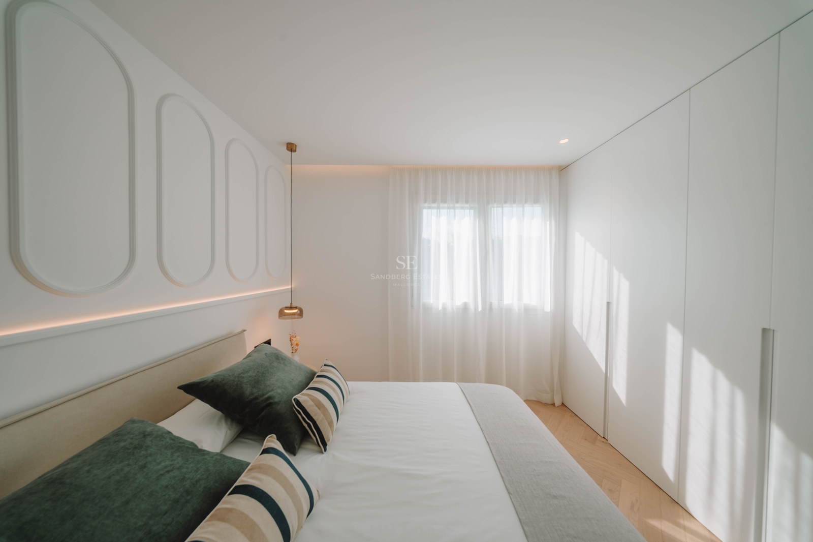 Bright bedroom with white paneled walls, a double bed with green pillows, herringbone wood floors, and built-in closets.