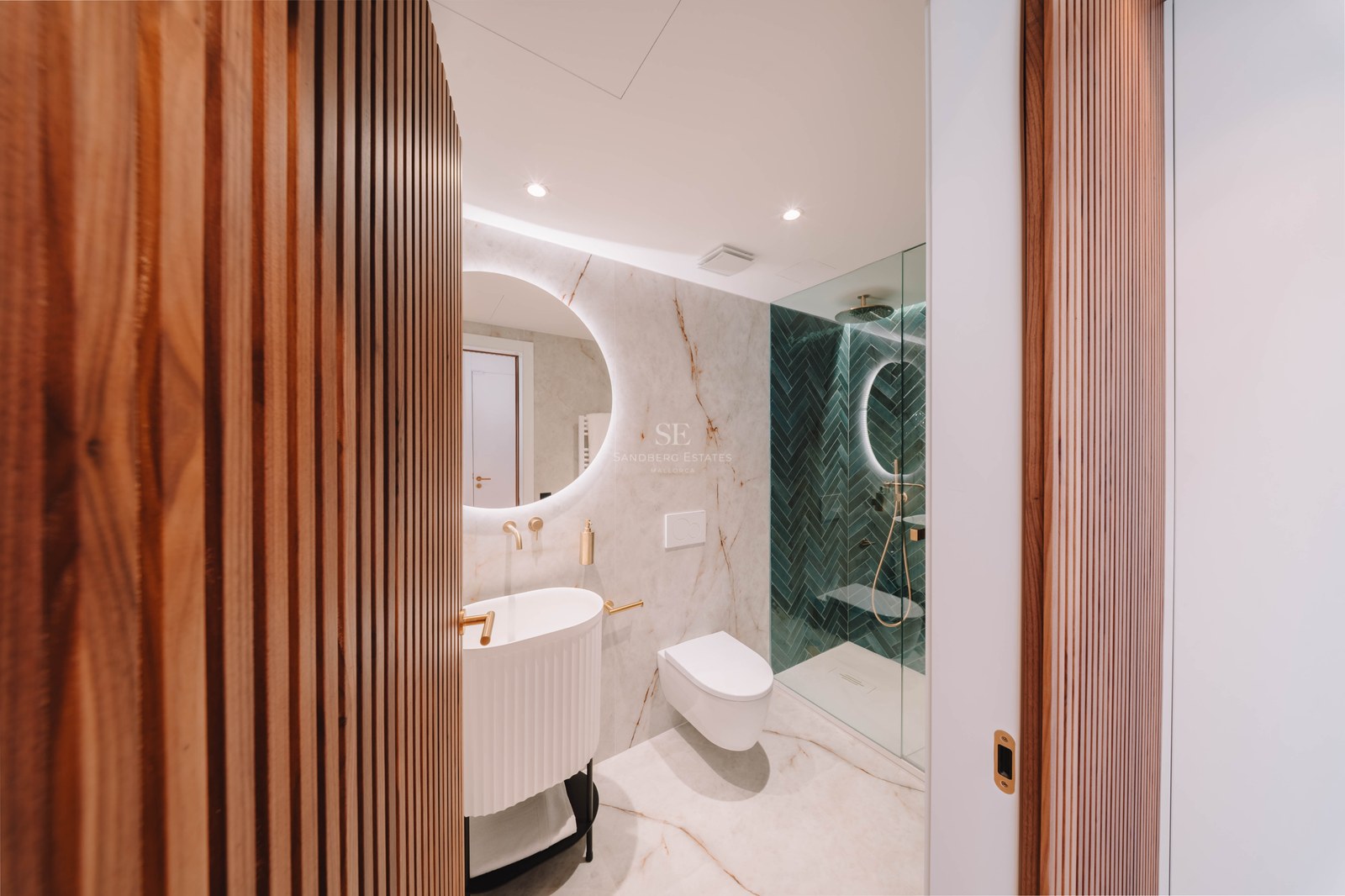 Modern bathroom featuring marble walls, a backlit circular mirror, gold fixtures, and green herringbone shower tiles.