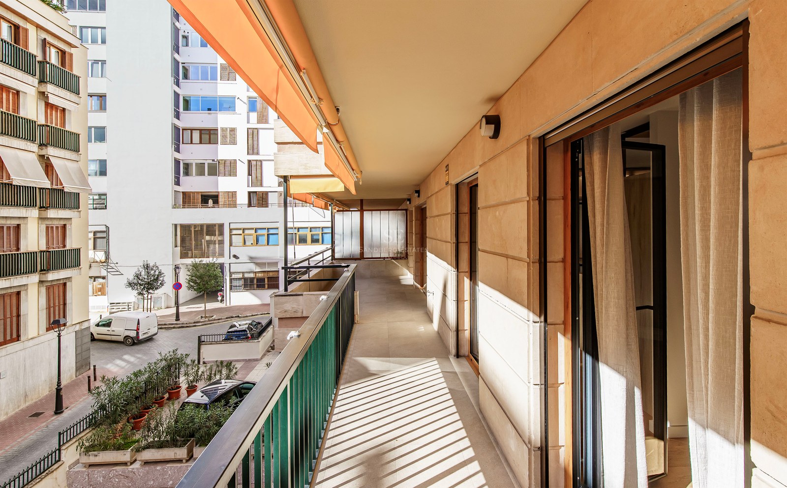 Long sunny balcony with orange awning and metal railing overlooking an urban street.