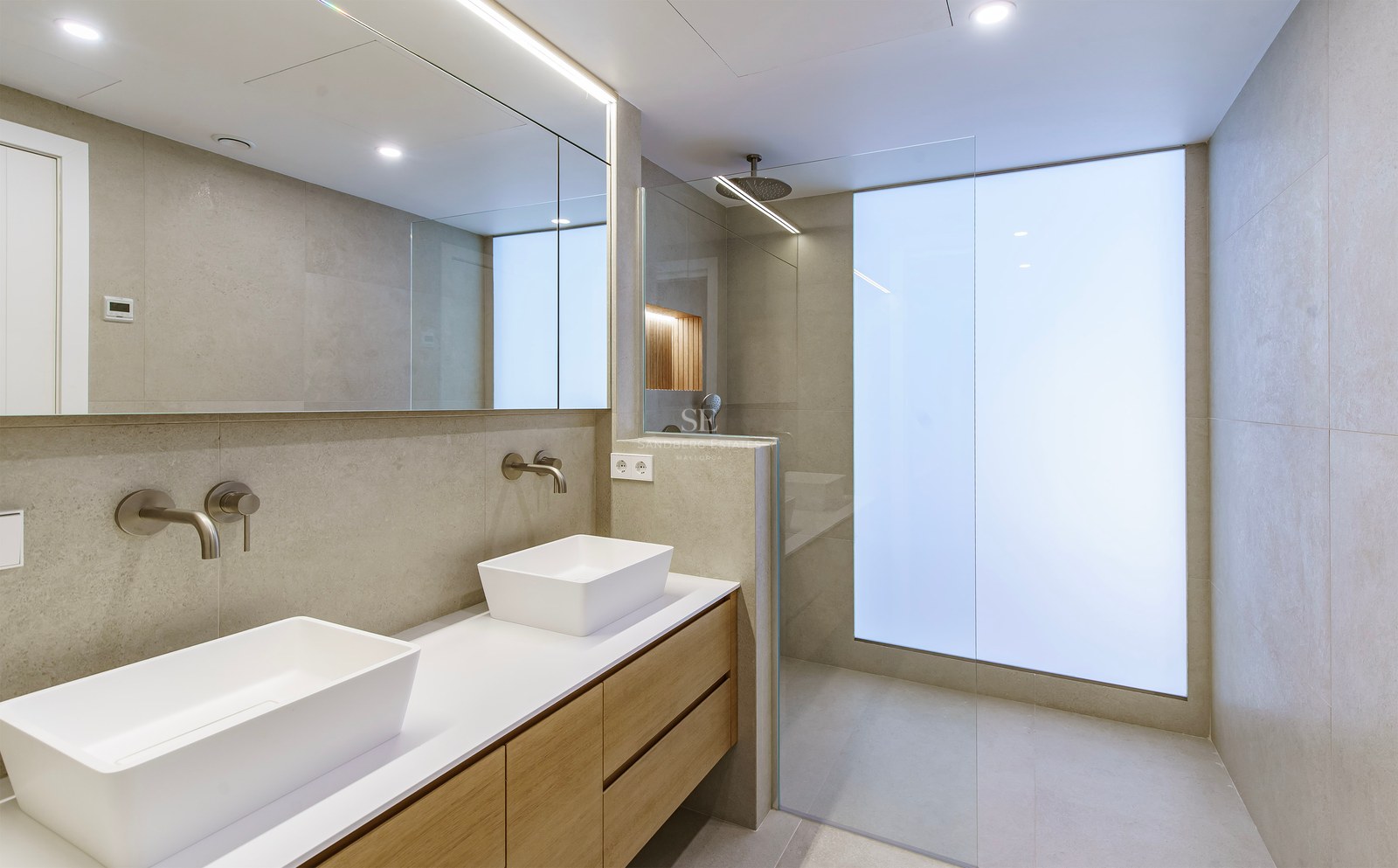 Minimalist bathroom with double white sinks on a wooden vanity, walk-in shower, and large backlit mirror.