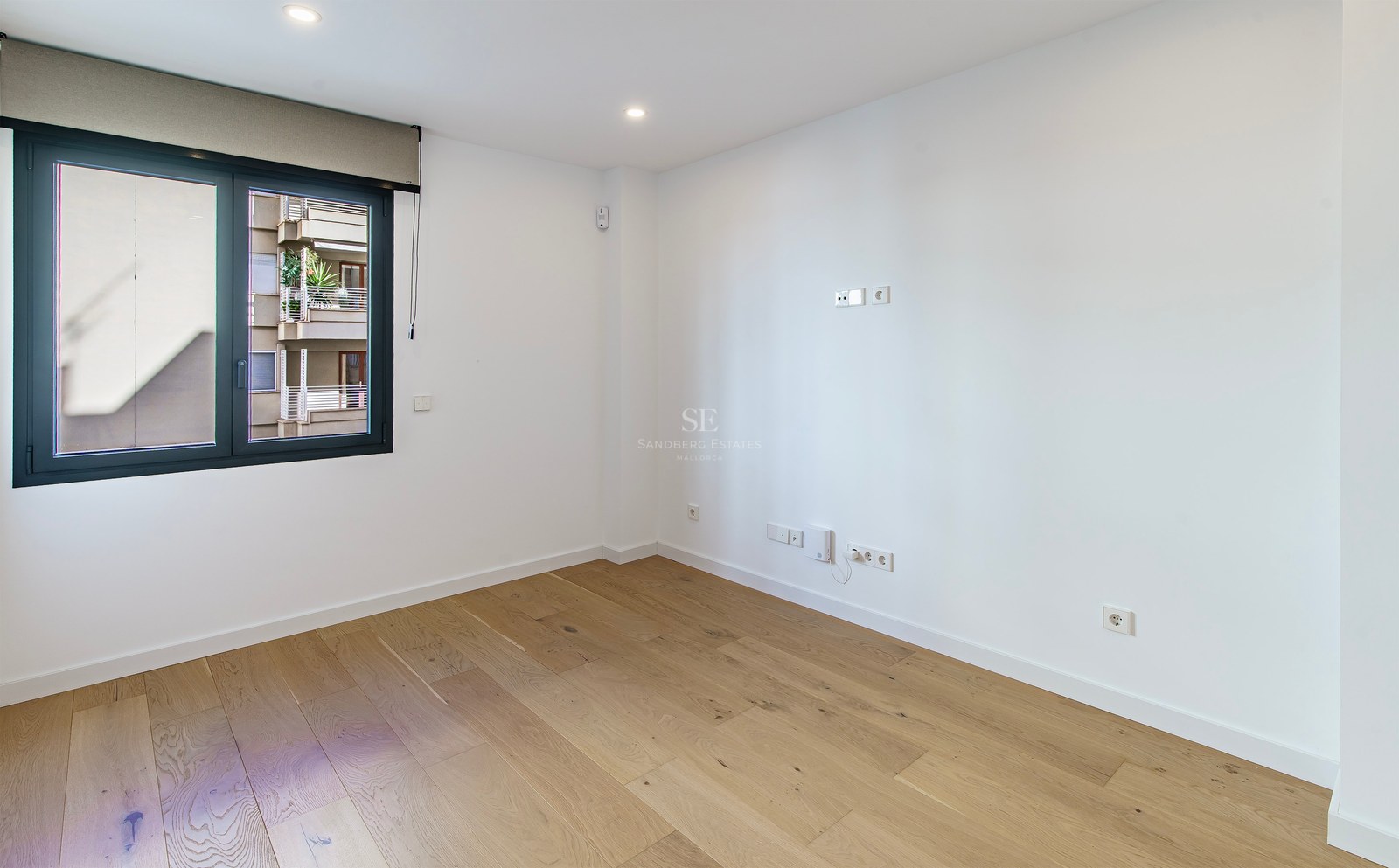 Bright empty room with light wood floors, white walls, and a large window with a roller blind.