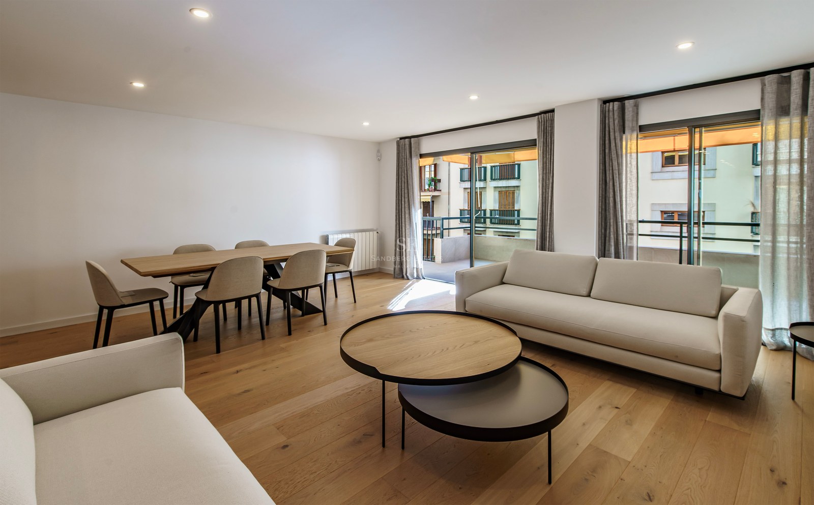 Modern living room featuring white sofas, wooden coffee tables, a dining set, and large windows leading to a balcony.