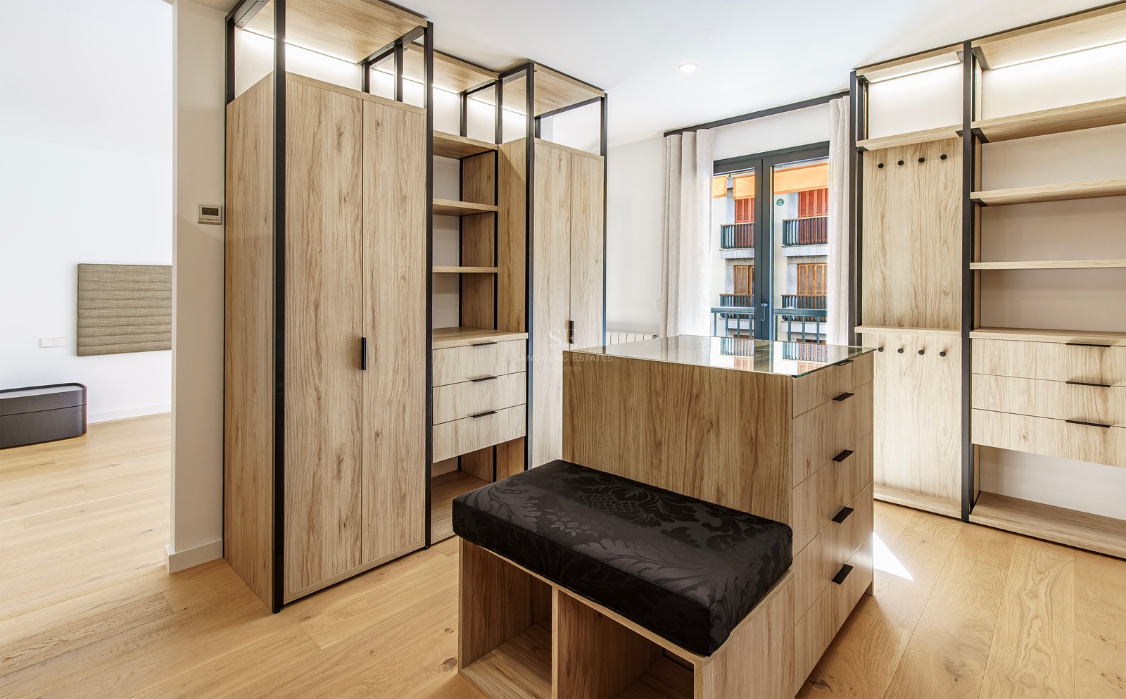 Spacious walk-in closet featuring light wood cabinetry, a central island with a glass top, and a black cushioned bench.