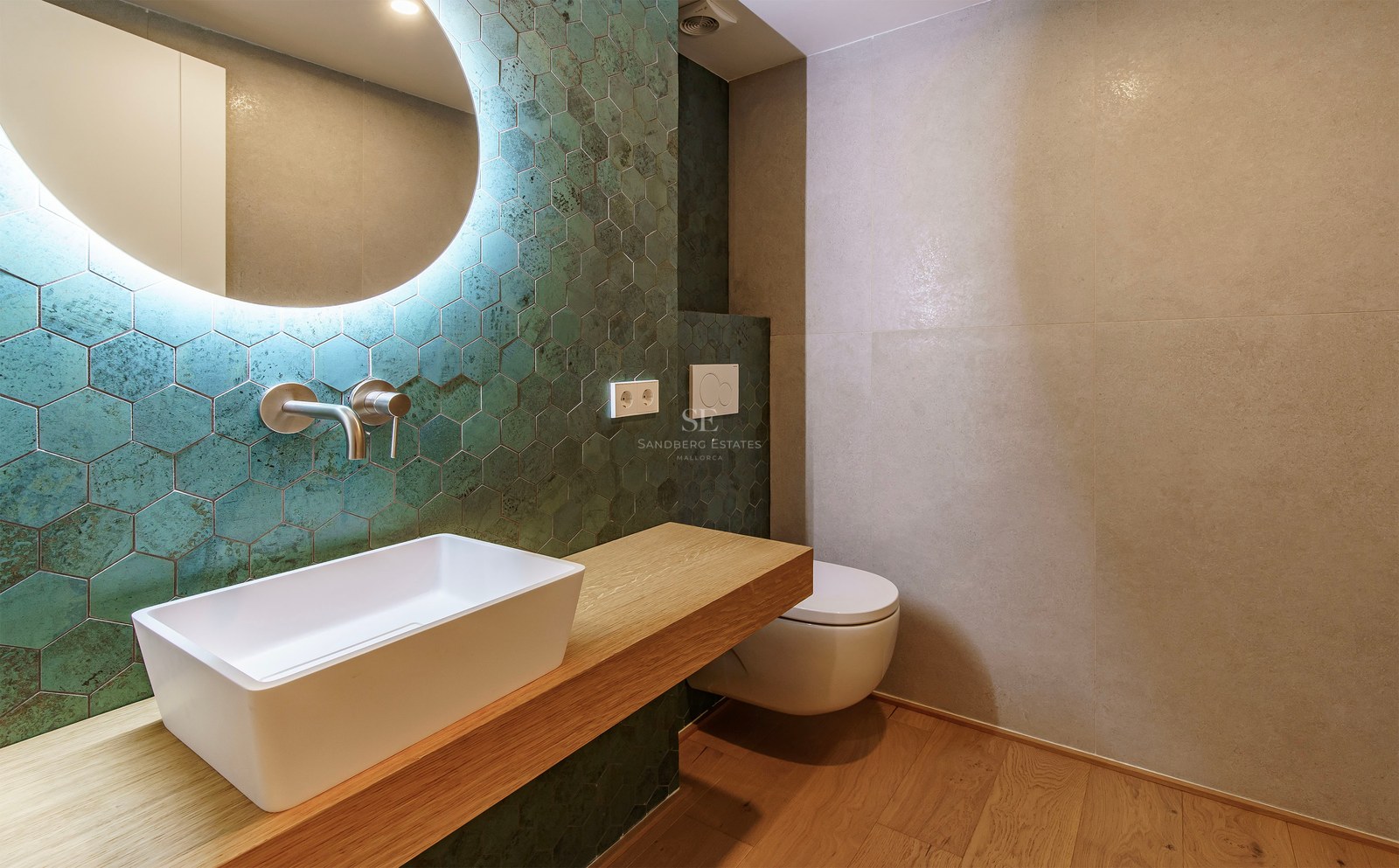 Modern bathroom featuring teal hexagonal tiles, a white vessel sink on a wooden vanity, and a large circular backlit mirror.