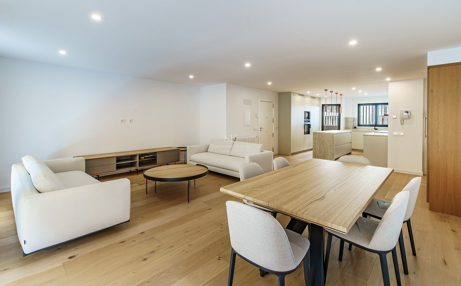 Spacious modern room featuring white sofas, a large wooden dining table, and a contemporary kitchen in the background.
