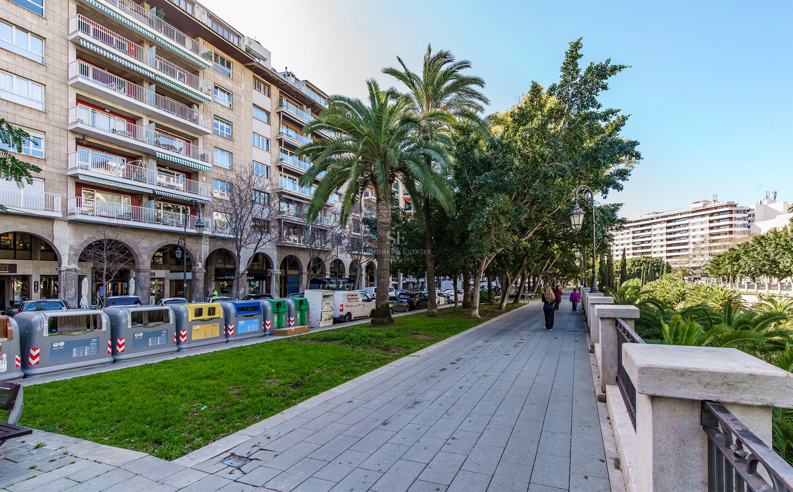 Paved city promenade with palm trees next to a multi-story stone building with balconies under a blue sky.