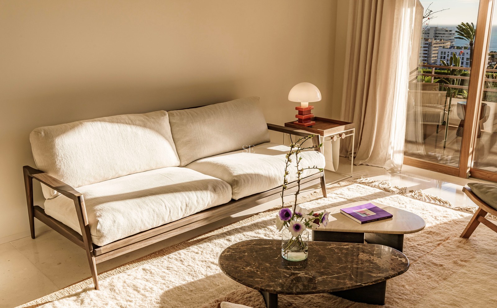 Cremefarbenes Sofa und Marmortische in einem sonnigen Wohnzimmer mit Balkon und Meerblick.