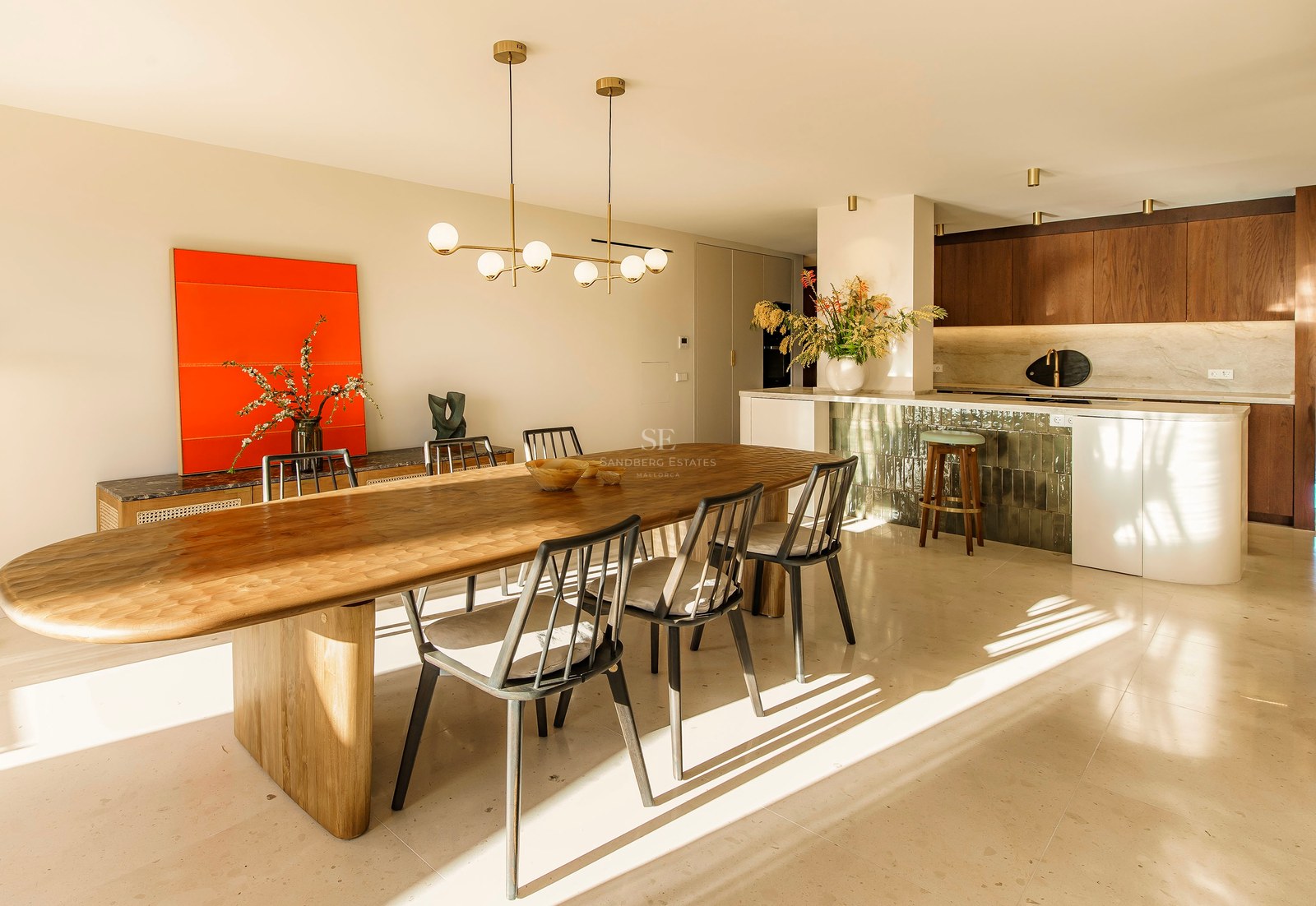 Großzügiges Esszimmer mit ovalem Holztisch, Designer-Pendelleuchte und moderner Küche im Hintergrund bei Sonnenlicht.
