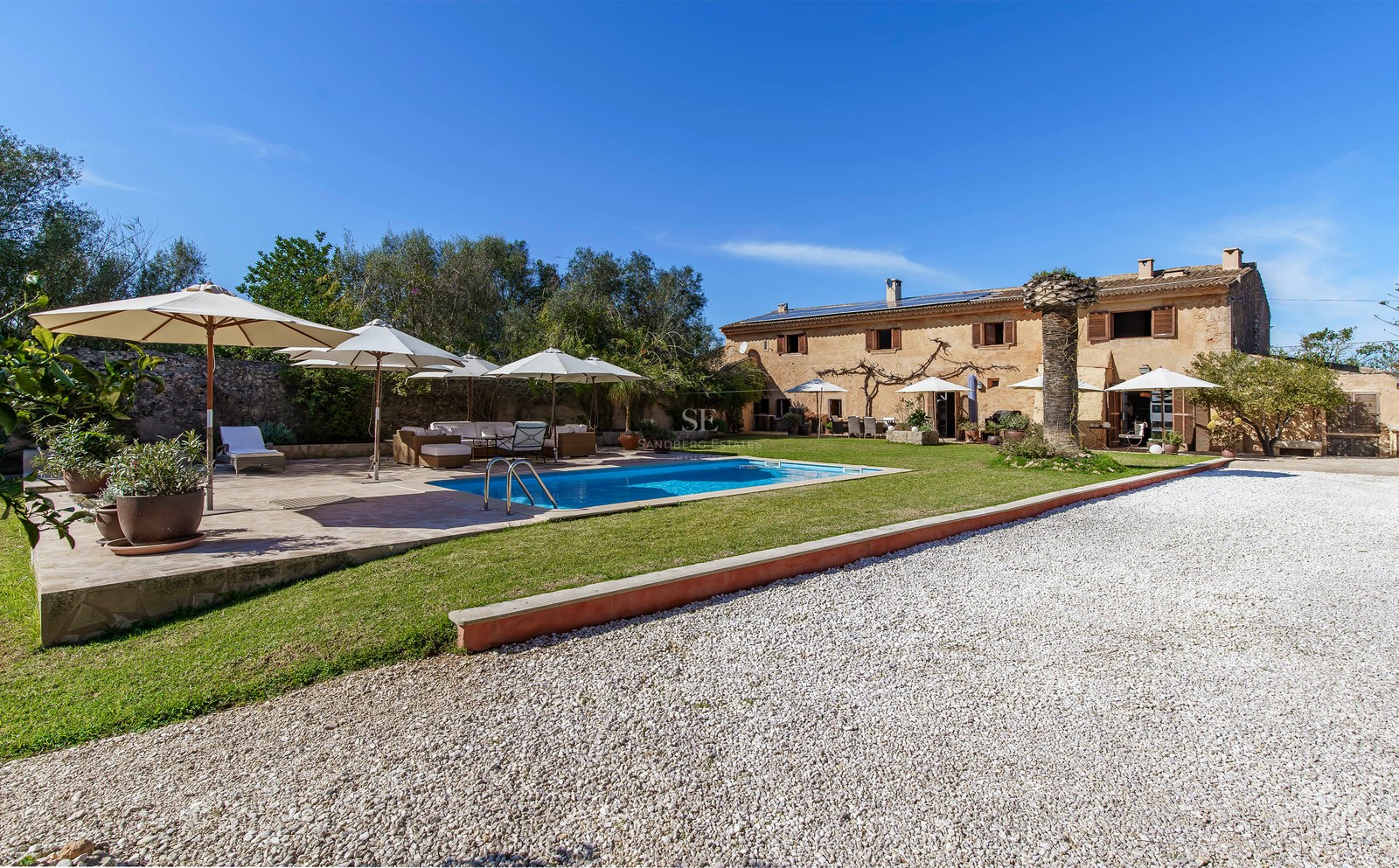Große Steinvilla mit blauem Pool, weißen Sonnenschirmen und Garten unter klarem Himmel.