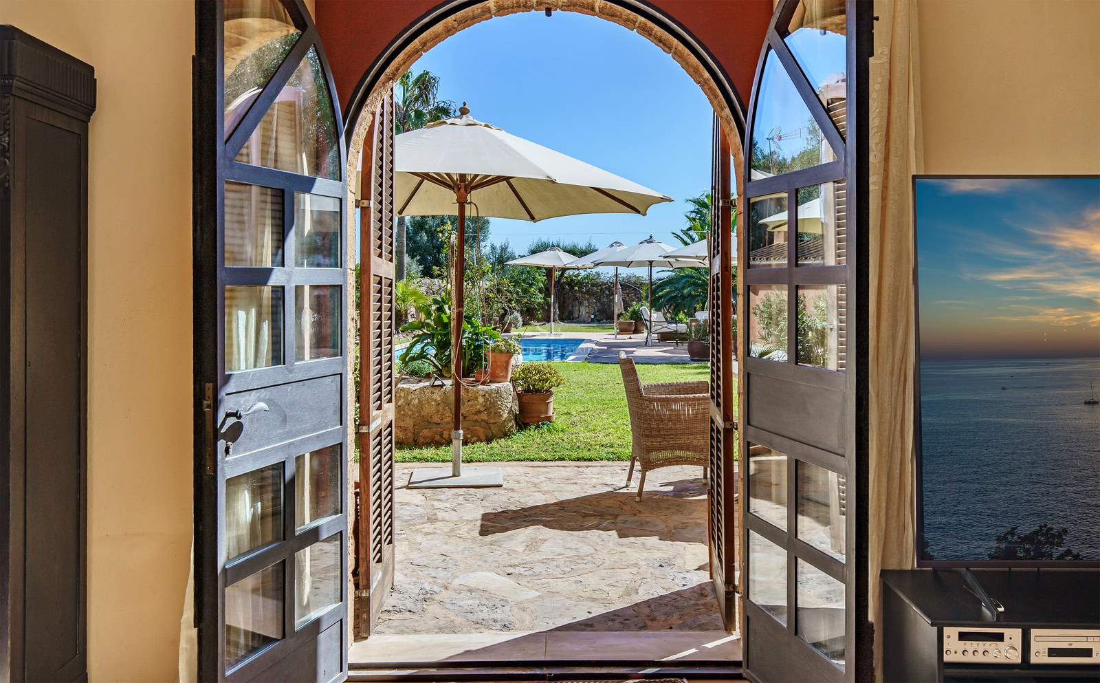 Blick durch offene Rundbogentüren auf einen privaten Pool, Garten und weiße Sonnenschirme unter blauem Himmel.