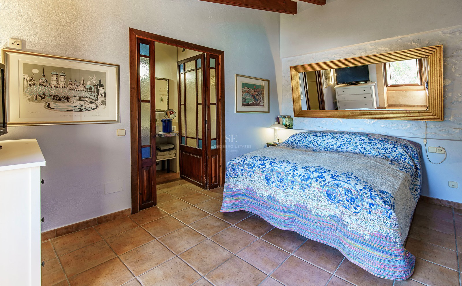 Schlafzimmer mit blau gemusterter Decke, Terrakottaboden, großem Goldspiegel und Holztüren zum Bad.