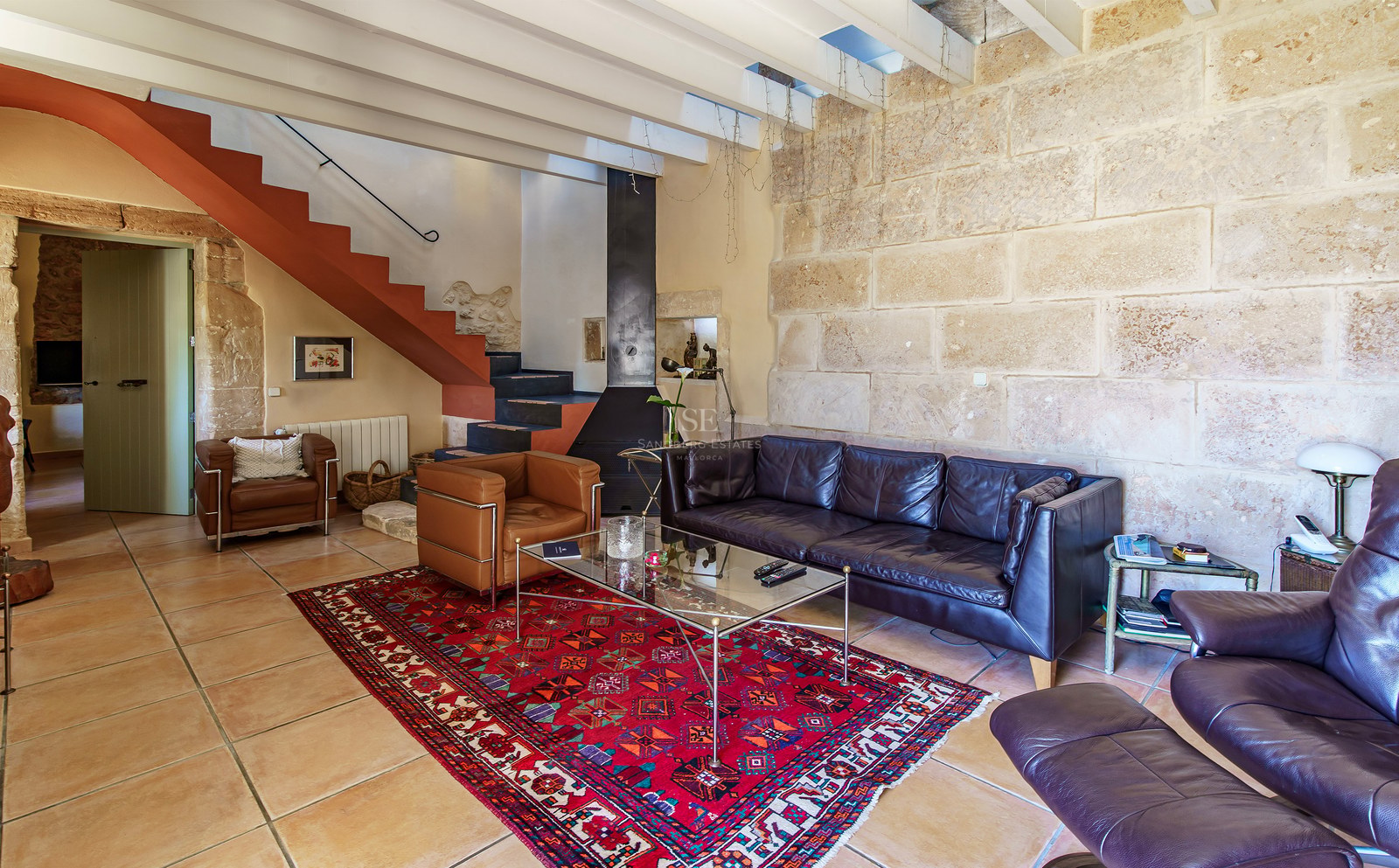 Wohnzimmer mit Ledersofas, rotem Teppich, Steinwand und Treppe mit Terrakotta-Akzenten.